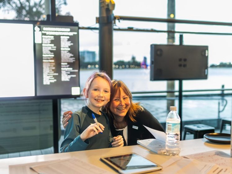 Image of A smiling adult and child sit together at a table with papers, a tablet, a water bottle, and hand sanitiser, in a bright room with large windows overlooking water.