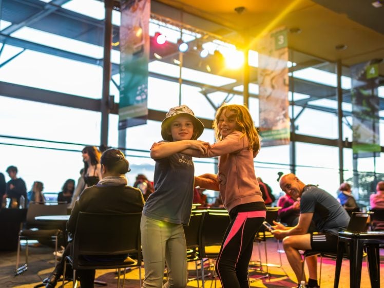 Image of Two children are smiling and dancing together in a bright, modern indoor space with large windows. People are seated and chatting in the background, with colourful lights shining from above.