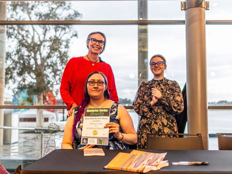 Image of Three women indoors, two standing and one seated at a table holding a certificate. The women are smiling, and there are books on the table. Large windows and trees are visible in the background.