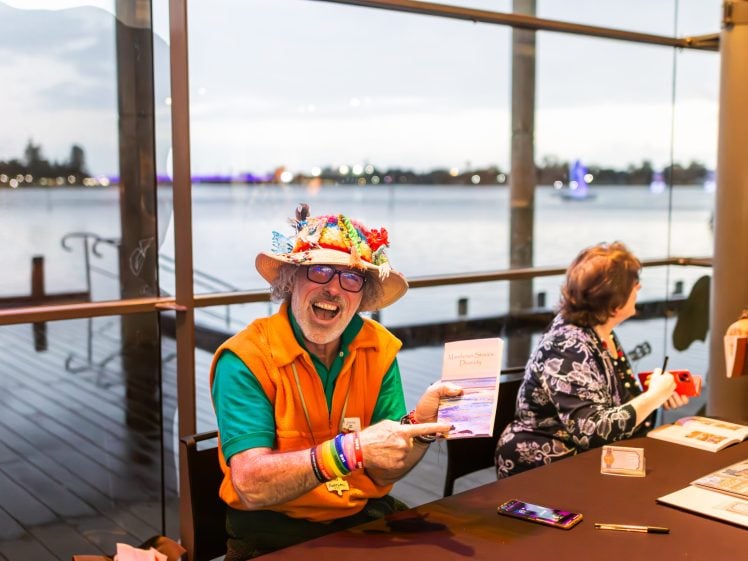 Image of A cheerful man in a colourful hat and orange waistcoat sits at a table by large windows overlooking a waterfront, smiling and holding up a booklet. Other people sit beside him, and event materials are on the table.