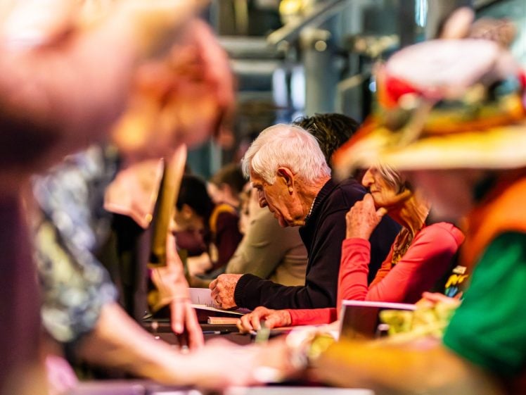 Image of Elderly people sit at a long table indoors, focusing on papers or forms. The image is vibrant with colourful clothing and hats, and shows a busy, lively atmosphere with some people blurred in the foreground.