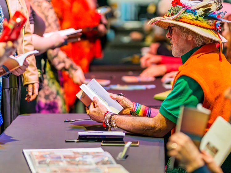 Image of A colourfully dressed person wearing a decorated hat sits at a table signing books for a queue of people at a lively indoor event. Other people are waiting, holding books and phones, as the person signs copies.