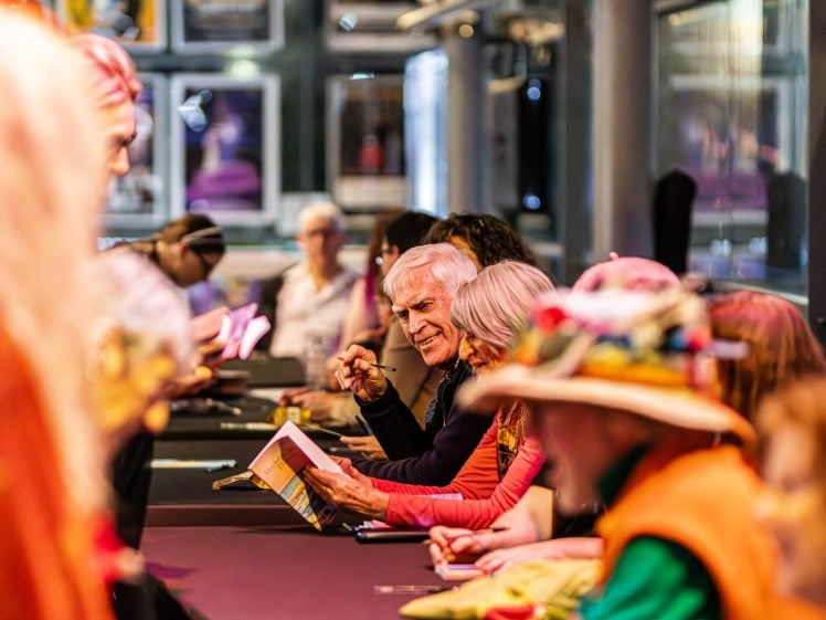 Image of A group of people sit at a long table indoors, signing books for attendees. One older man in focus is smiling and interacting with someone across the table. The scene is colourful and lively, with blurred figures in the foreground.