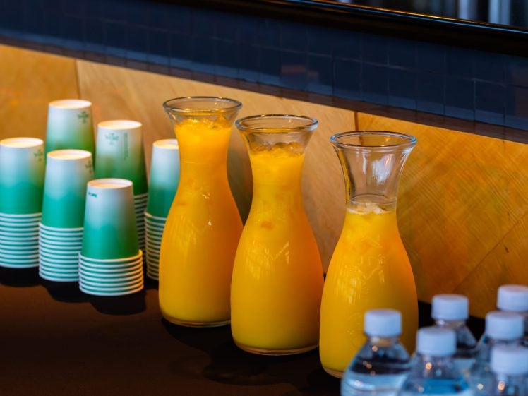 Image of Three glass carafes filled with orange juice are placed on a dark table beside stacked green and white paper cups and bottled water. The background features a wooden and tiled wall.