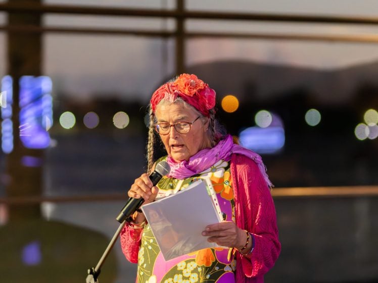 Image of An older woman with grey hair, wearing glasses, a pink scarf, and a colourful floral dress, reads from papers while speaking into a microphone. She stands indoors with blurred lights in the background.