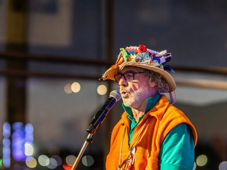 Image of An older man wearing glasses, an orange waistcoat, and a colourfully decorated hat sings or speaks into a microphone indoors under soft lighting, with blurred lights visible in the background.