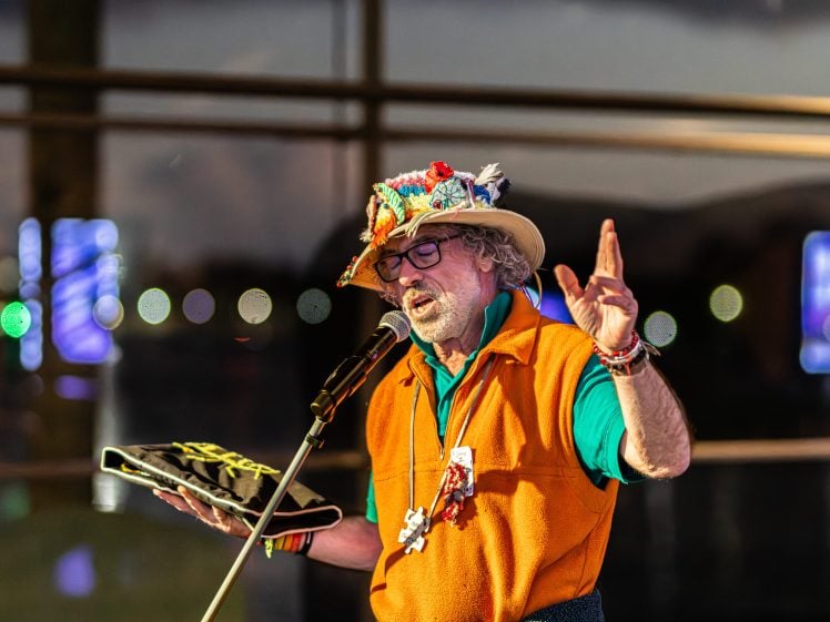 Image of A man in a colourful hat and orange waistcoat speaks or sings passionately into a microphone, holding a black item in one hand, with blurred lights and windows in the background.