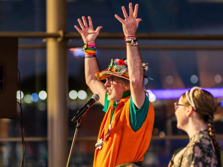 Image of A person wearing a colourful hat, orange waistcoat, and multiple bracelets raises both hands whilst speaking or singing into a microphone. Another person with glasses looks on in the foreground. Bright lights blur in the background.