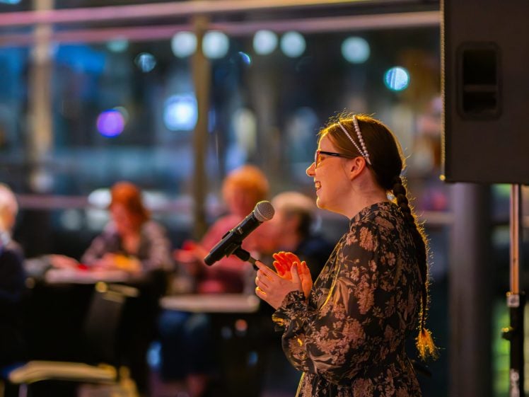 Image of A woman with glasses and a patterned dress stands at a microphone, smiling as she speaks to an audience seated at tables in a softly lit, modern indoor setting.