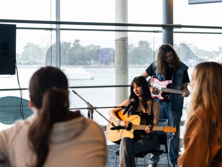 Image of A woman plays an acoustic guitar and sings into a microphone, whilst a man behind her plays an electric guitar. They perform indoors near large windows with an audience watching.