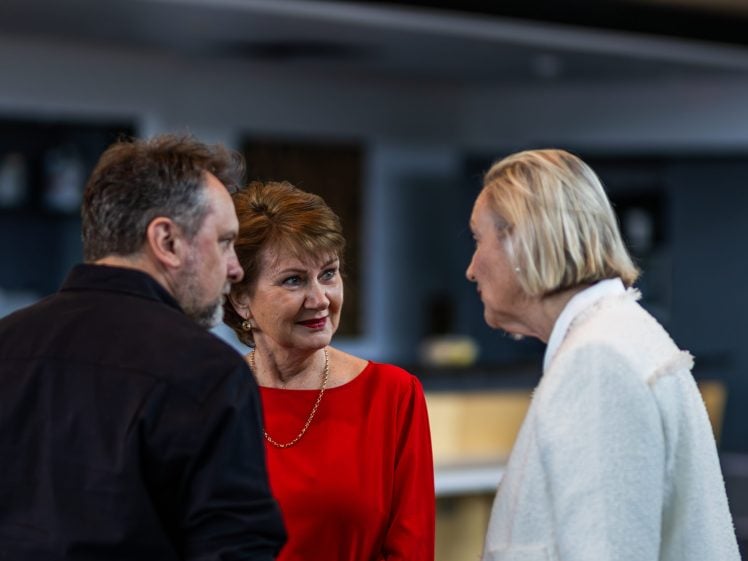 Image of Three adults are standing together indoors, engaged in conversation. One man faces two women; one woman wears a red dress and smiles, while the other wears a white jacket. The background is softly blurred.