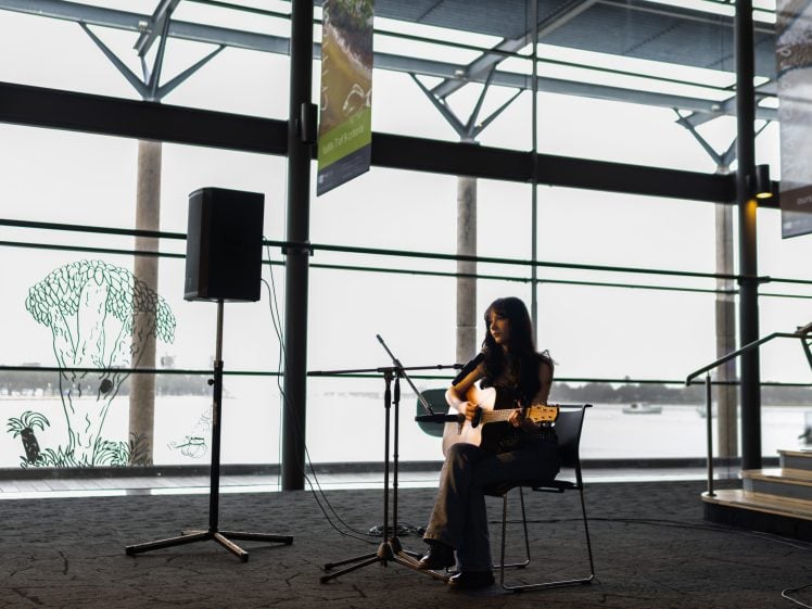 Image of A woman sits on a chair indoors, playing an acoustic guitar and singing into a microphone. She is silhouetted against large windows with a waterfront view, and audio equipment is set up nearby.