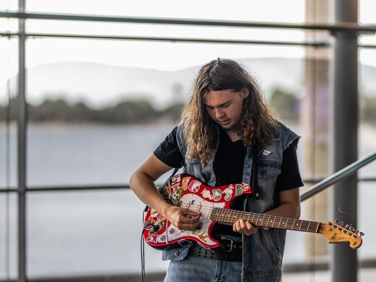 Image of A young man with long hair plays an electric guitar decorated with red and white stickers, standing indoors by large windows with an outdoor view in the background.