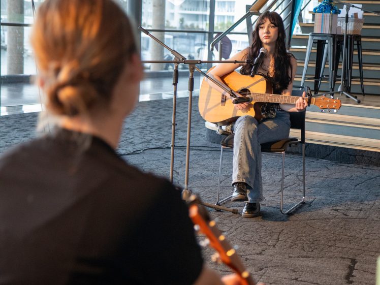 Image of A young woman sits and plays an acoustic guitar, singing into a microphone. Another person with a guitar is in the foreground, facing her. They are indoors near large windows and a staircase.