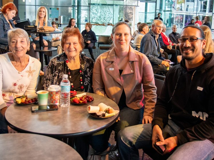 Image of Four people, three women and one man, sit around a table with food and drinks at a busy indoor café. Other patrons are visible in the background, and the atmosphere is lively and sociable.