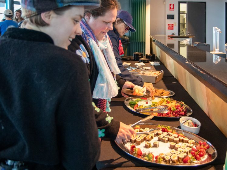Image of Three people serve themselves food from a buffet table with platters of assorted snacks, including fruit, desserts, and biscuits, in a modern indoor setting.