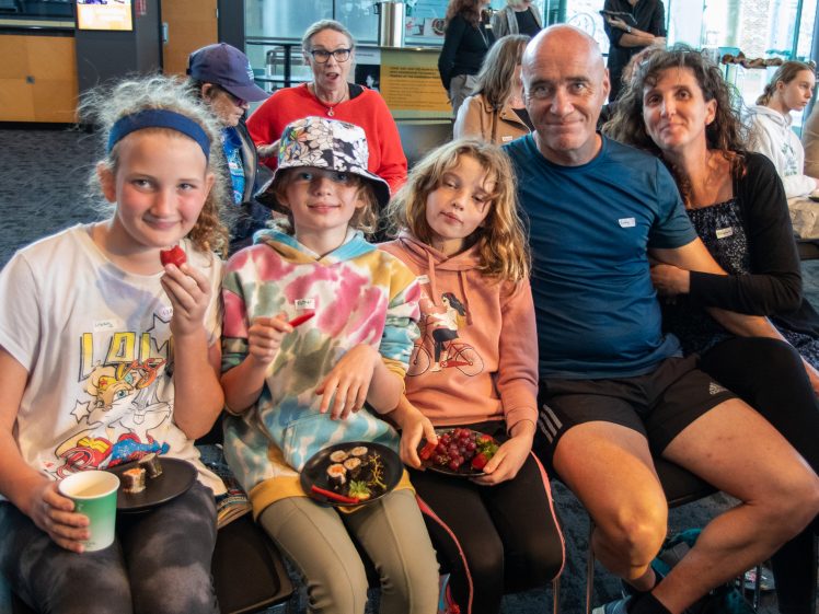 Image of A family of five, including three children and two adults, sit closely together indoors, smiling at the camera. The children hold plates with snacks, and people are visible in the background.