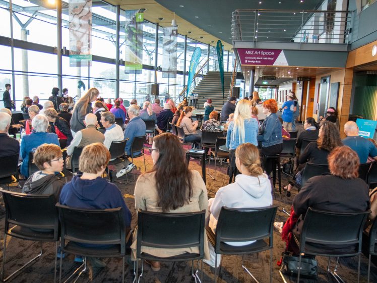 Image of A large group of people sit and stand in a modern, glass-walled event space with banners, tables, and a staircase visible. The atmosphere is lively and sociable, with natural light filling the room.