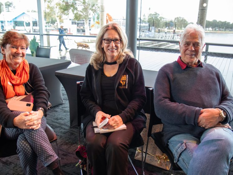 Image of Three older adults, two women and one man, sit indoors by large windows overlooking a waterfront. They are smiling and appear relaxed, with the women holding a notebook and a tablet.