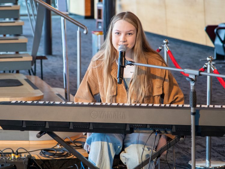 Image of A young woman with long blonde hair sits at a Yamaha keyboard, singing into a microphone. She wears a brown jacket and jeans, performing indoors near red rope barriers and a staircase.