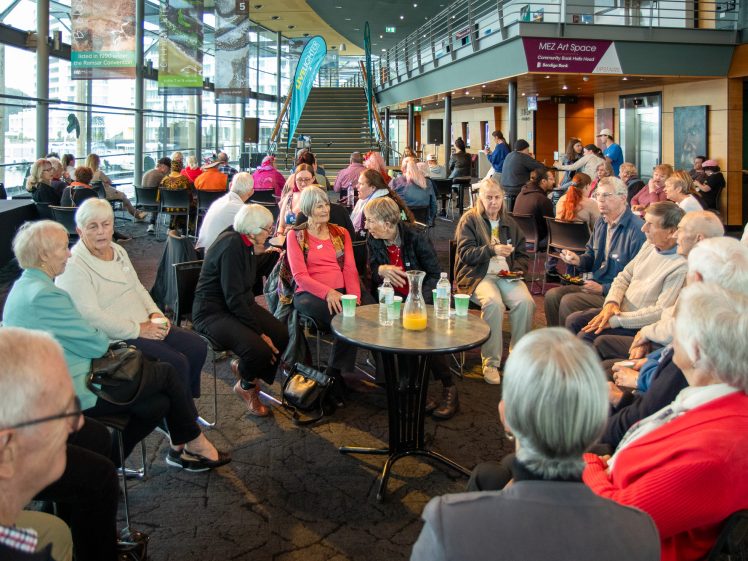 Image of A large group of older adults sit in a circle at round tables in a modern indoor venue, chatting and enjoying drinks. The space is bright with glass walls, and others are visible in the background.