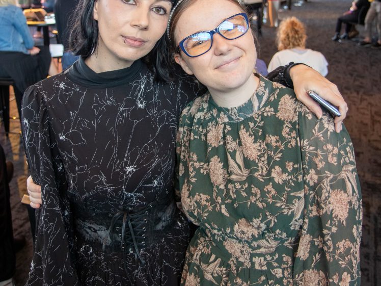 Image of Two women stand close together indoors, smiling at the camera. One has dark hair and wears a black patterned dress; the other wears glasses, a headband, and a green floral dress. Other people are visible in the background.