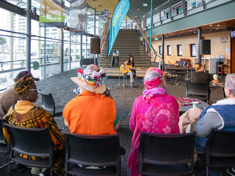 Image of A group of people in colourful clothing sit facing a musician playing guitar on a small stage in a modern, well-lit indoor venue with large windows and a staircase in the background.