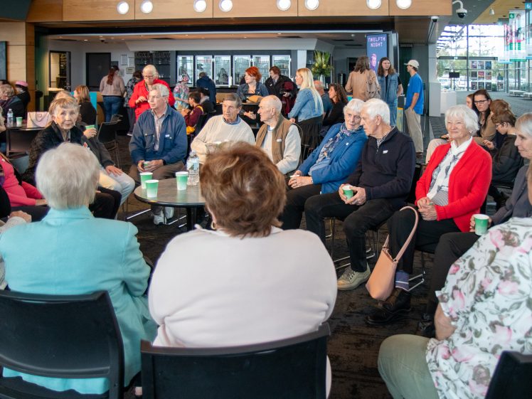 Image of A group of older adults sit in a circle at an indoor community event, engaging in conversation. Other people mingle in the background. The setting is bright with large windows and modern décor.