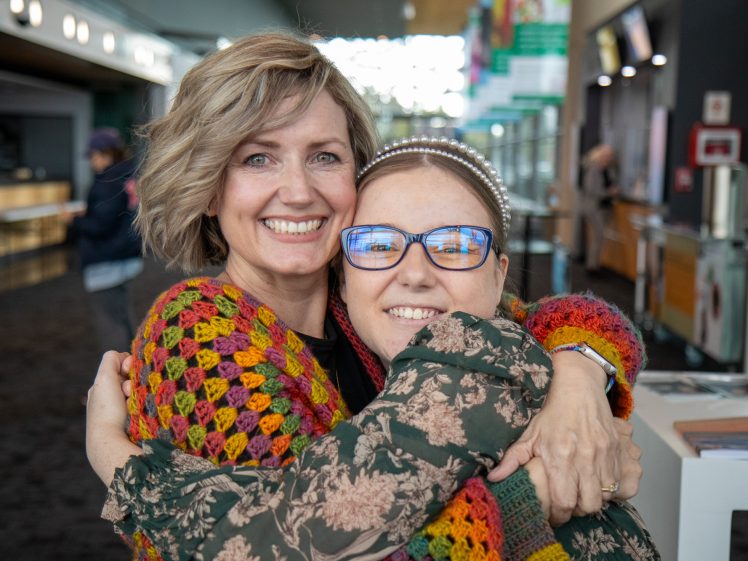 Image of Two women smiling and hugging each other warmly indoors, both wearing colourful clothing. One has glasses and a headband; the other has short, wavy hair. The background shows a modern, well-lit public space.