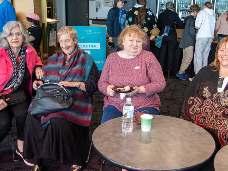 Image of Four older women sit at two round tables, smiling at the camera. In the background, people interact near a refreshments area with a teal sign and cups. The setting appears to be a casual indoor event or gathering.