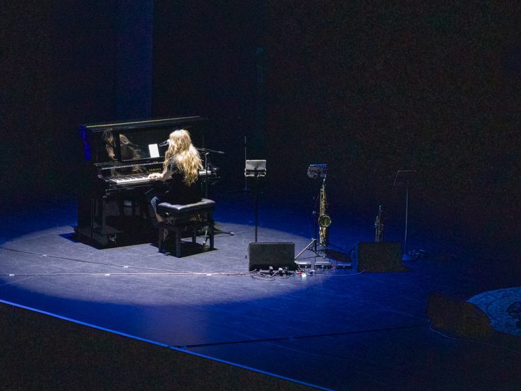 Image of A woman with long hair plays a grand piano on a dark stage, illuminated by a spotlight. Nearby, a saxophone and microphone stand are visible, with cables and equipment on the floor.