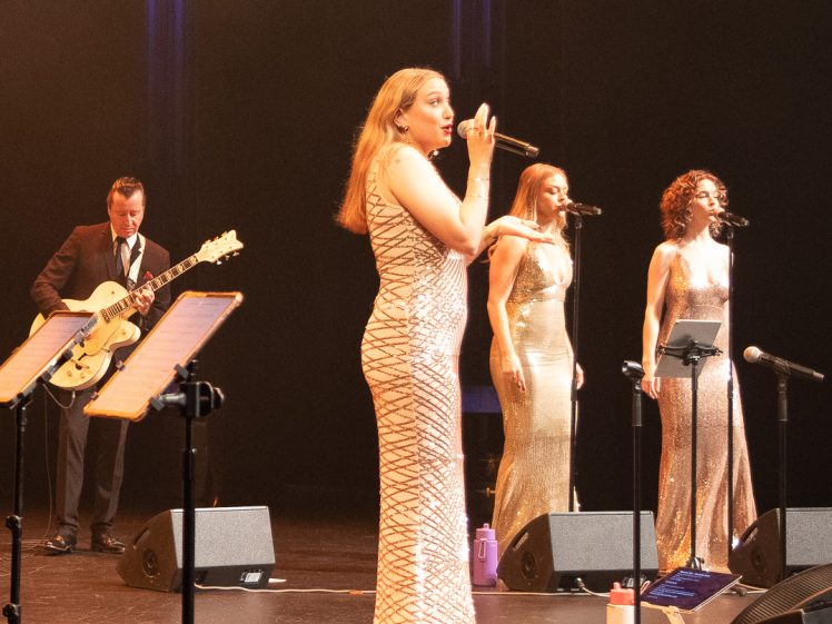 Image of Three women in shimmering dresses sing on stage with microphones, while a man in a suit plays an electric guitar. Music stands and water bottles are visible, and the stage is dimly lit with spotlights.