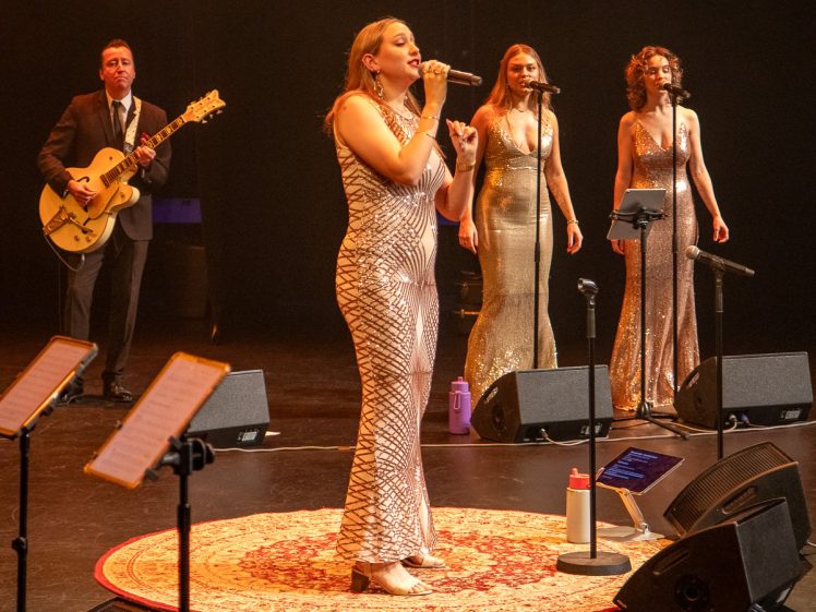 Image of A female singer in a sparkling gown performs on stage with two backing singers in similar dresses and a guitarist in a suit. They stand on a lit stage with microphones and music stands, and a patterned rug under the lead singer.