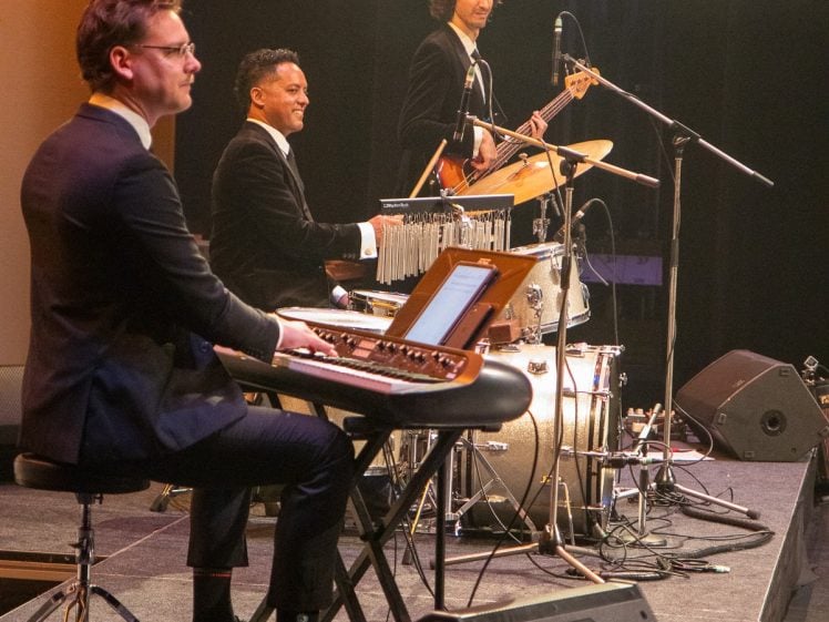 Image of Three musicians in suits perform on stage; one plays a keyboard, another is on drums, and the third plays bass guitar. They are focused on their instruments, surrounded by microphones and stage equipment.