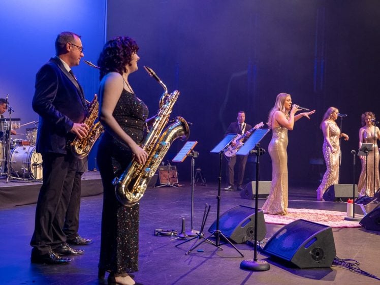 Image of A band performs on stage with musicians playing saxophones and drums while three women in gold sequin dresses sing into microphones. The stage is lit with blue and purple lighting.