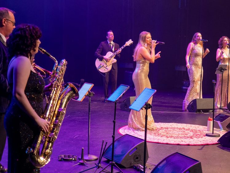 Image of A band performs on stage. Three women in gold dresses sing into microphones, a man in a suit plays guitar, and another woman in a dark dress plays saxophone. Stage lights create a colourful atmosphere.