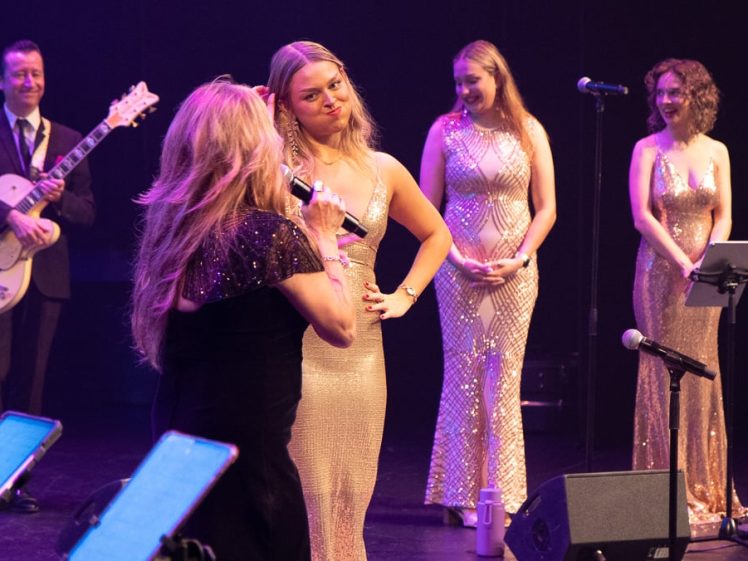 Image of A group of performers on stage, including a guitarist and four women in elegant, shimmering dresses. One woman sings into a microphone whilst another stands facing her with hands on hips. The scene is brightly lit.