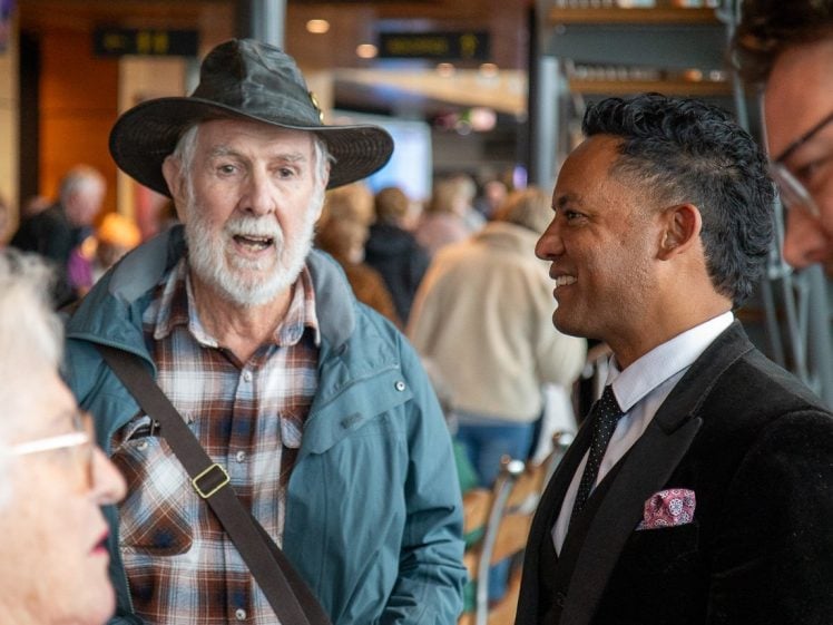 Image of Two men stand and converse at an indoor event. One wears a hat, checked shirt, and teal jacket, while the other is dressed in a black suit and tie. People and blurred activity fill the background.