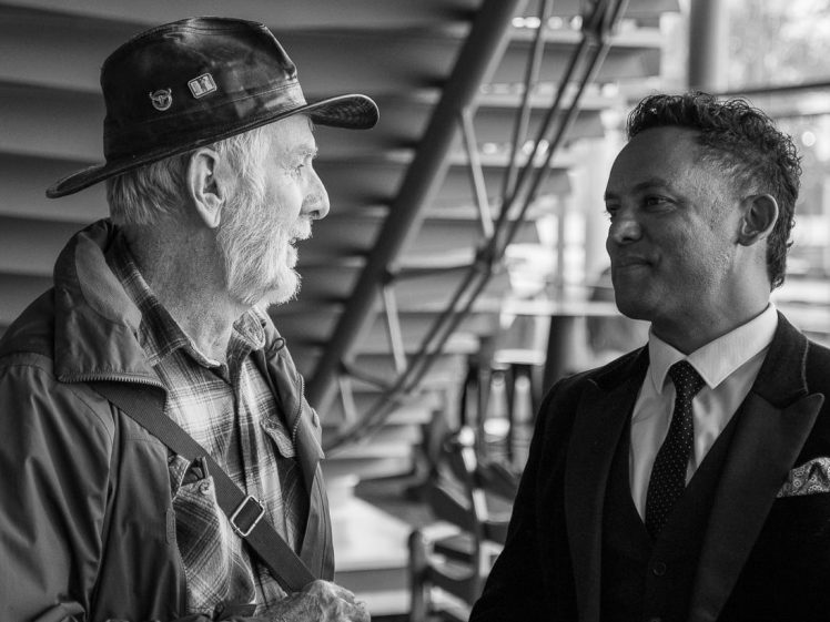 Image of Two men stand facing each other and smiling. One is older, wearing a hat and casual jacket, while the other is younger, dressed in a suit and tie. They appear to be having a friendly conversation indoors.