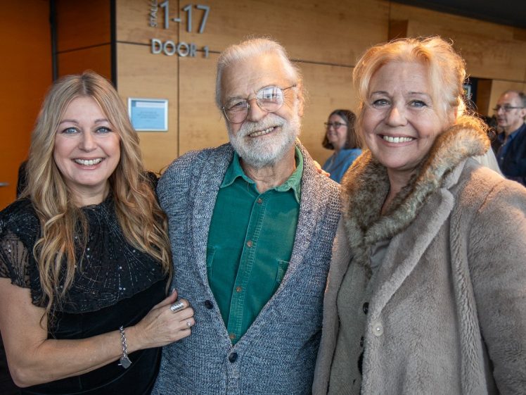 Image of Three smiling adults, two women and one man, pose together indoors. The man wears glasses, a green shirt, and a grey cardigan. One woman wears a black dress and the other a beige coat with a fur collar. Other people are seen in the background.