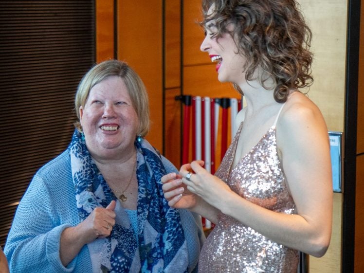Image of Two women smile and laugh together indoors; one wears a light blue scarf and jumper, giving a thumbs up, while the other wears a sparkly sleeveless dress. Warm light and colourful stripes are visible in the background.
