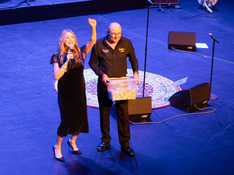 Image of A woman in a black dress speaks into a microphone and raises her arm, while a man in black holds a clear box filled with tickets or slips, standing on a stage with microphones and a round rug.