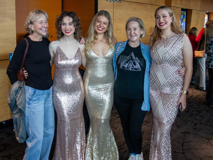 Image of Five women stand together, smiling for a group photo indoors. Three of them wear sparkling gowns, while the other two are in casual clothing. The background features wood-panelled walls and other people in the distance.