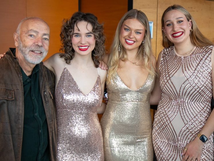 Image of Four people pose together indoors, smiling at the camera. Three women wear shimmery formal dresses in gold and champagne tones, while the man wears a brown jacket over a dark shirt. The background features warm wooden panelling.
