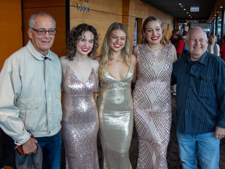 Image of Five people pose together indoors; three women in sparkly dresses stand between two older men, all smiling at the camera, in a wood-panelled venue with other people visible in the background.