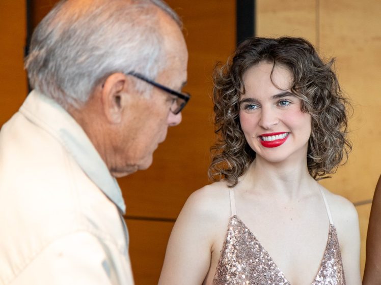 Image of A young woman in a sparkly, sleeveless dress smiles brightly at an older man wearing glasses, who is seen in profile. They appear to be having a friendly conversation indoors.