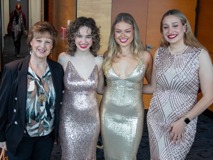 Image of Four women stand together indoors, smiling at the camera. Three are wearing sparkly dresses in shades of gold and beige, while the fourth, older woman wears a patterned blouse and black jacket.