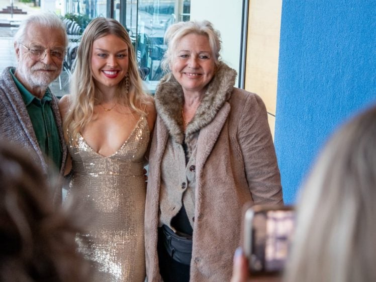 Image of Three people pose and smile together indoors, with one young woman in a sparkly gold dress standing between an older man in glasses and an older woman in a coat with a fur collar. A person in the foreground takes their photo.