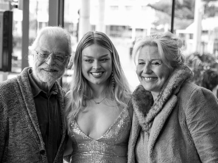 Image of Three people pose together, smiling for the camera. The young woman in the centre wears a sparkly dress, flanked by an elderly man in glasses and cardigan on the left and an elderly woman in a fur-trimmed coat on the right. The photo is in black and white.