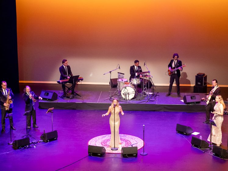 Image of A band performs on stage with a female singer in the centre. Musicians play saxophones, trumpet, keyboard, drums, guitar, and backing singers stand to the right. The background is lit with warm orange lighting.